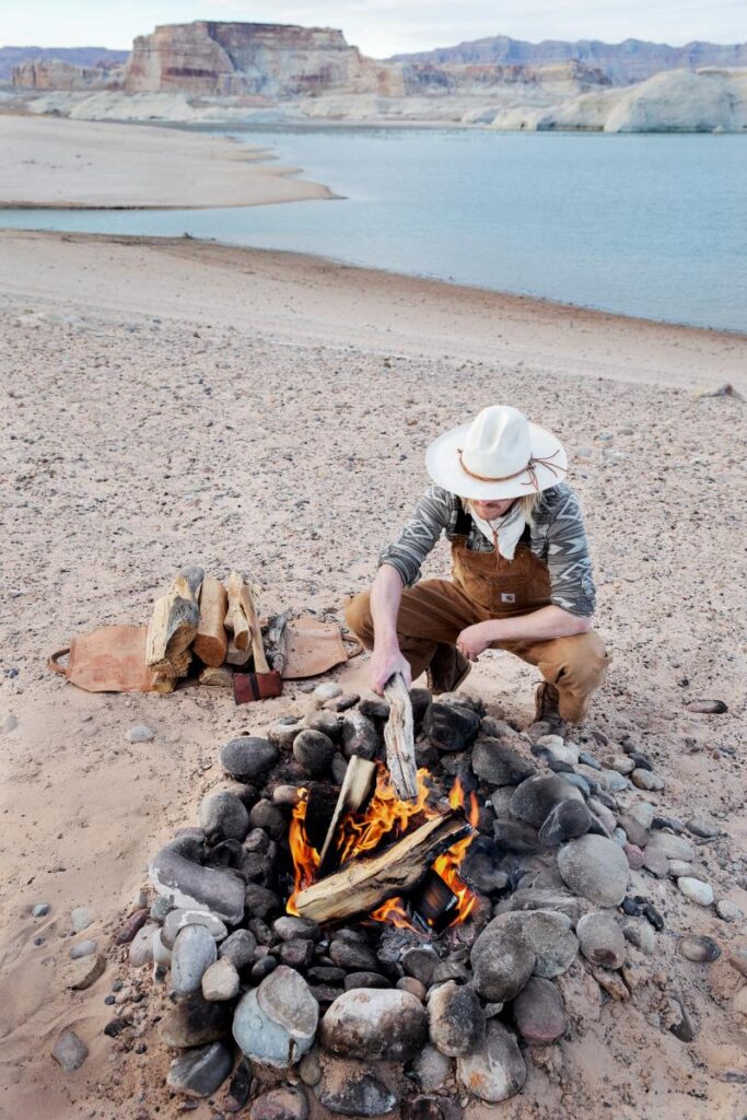 A person wearing a wide-brimmed hat and overalls kneels on a sandy beach, tending to a campfire with a metal poker.
