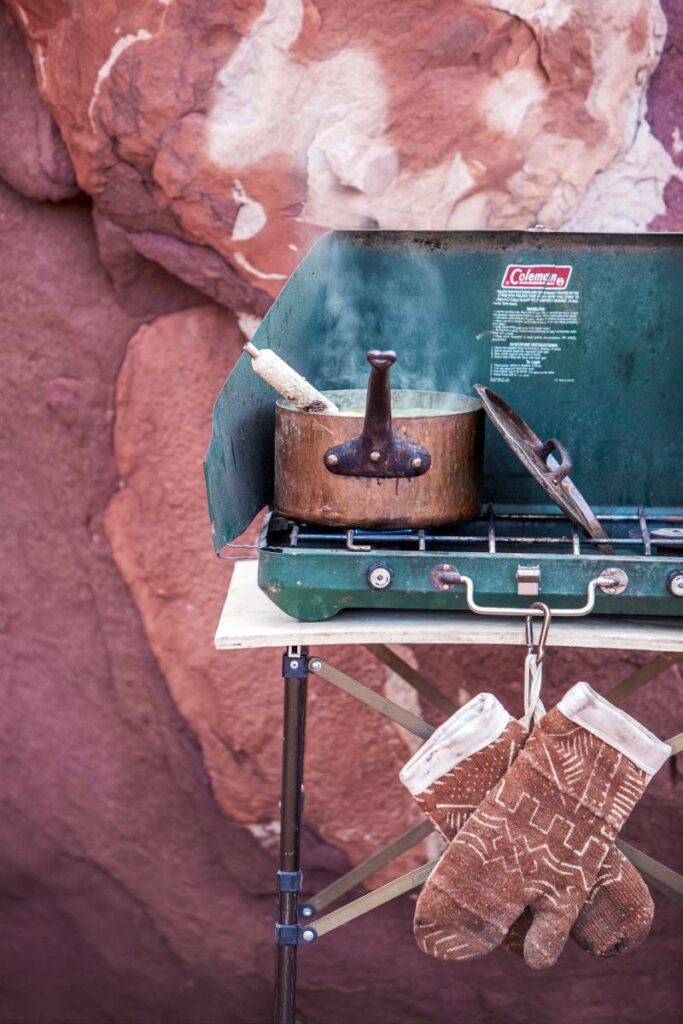 A copper pot with a lid sits on a green Coleman camping stove, with steam rising from it, set against a backdrop of reddish-brown rock formations.