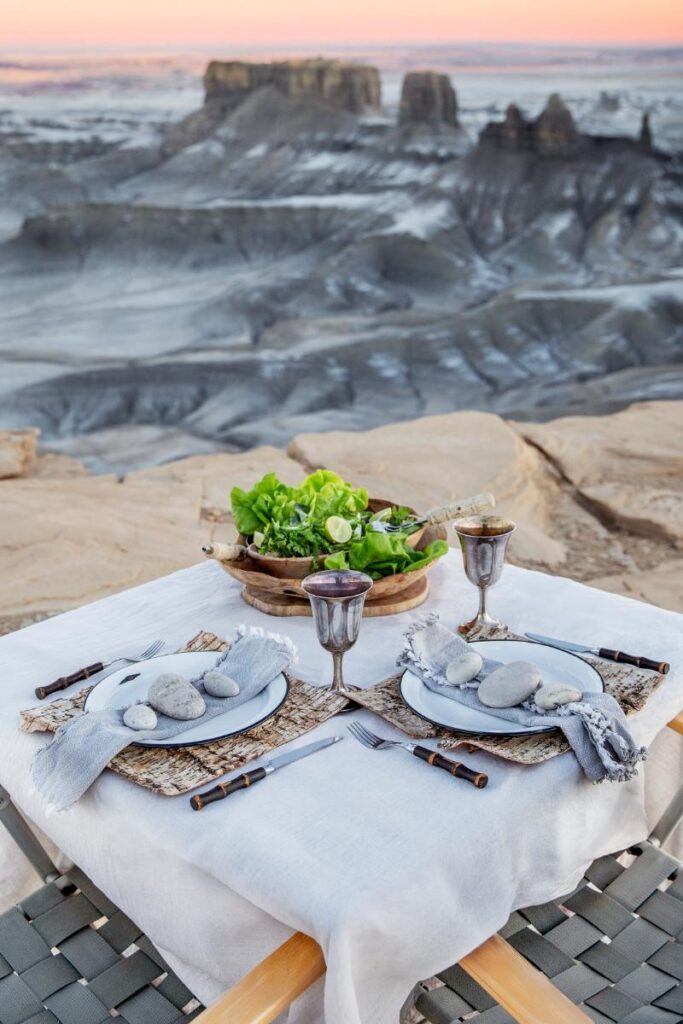 A set table for two in a desert landscape features white plates with light grey napkins and decorative stones, silver goblets, and a wooden bowl of salad.