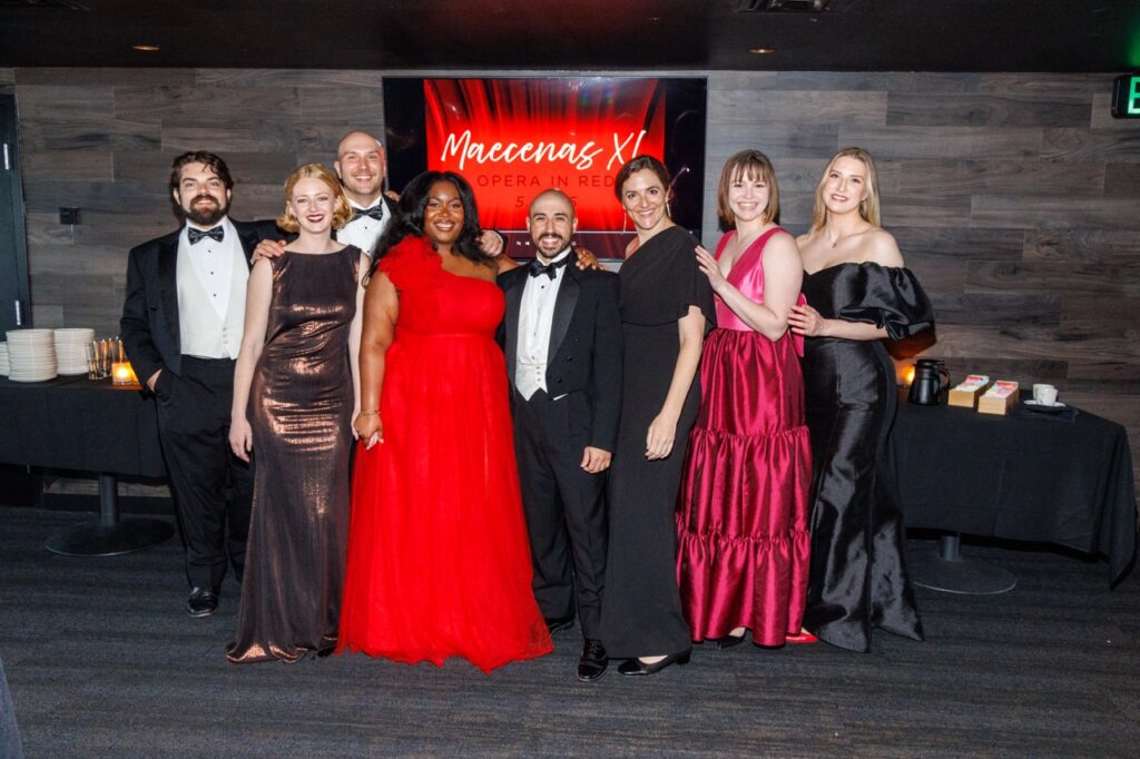 A group of men in suits and bowties and women in long dresses stand in front of black tables.