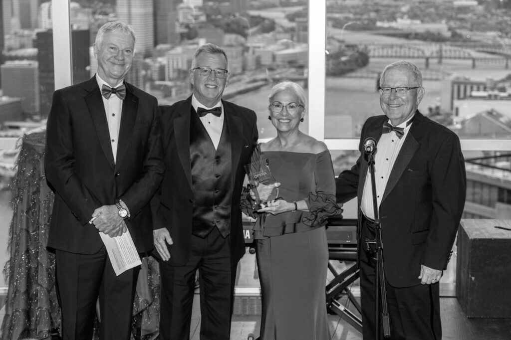 Three men in suits and bowties give an award to a woman in a lover dress and glasses.