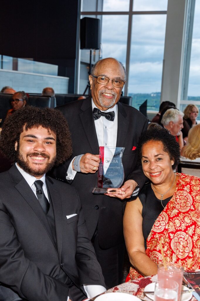A woman and a man sit at a table as another older man with glasses and a beard stands behind them with an award in his hands.