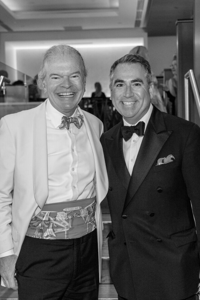 Two men in suit jackets and bowties smile together in a black and white photograph.