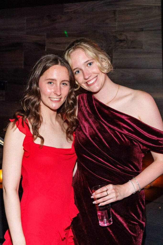 A woman in a bright red dress stands with a woman in a dark velvet red dress.