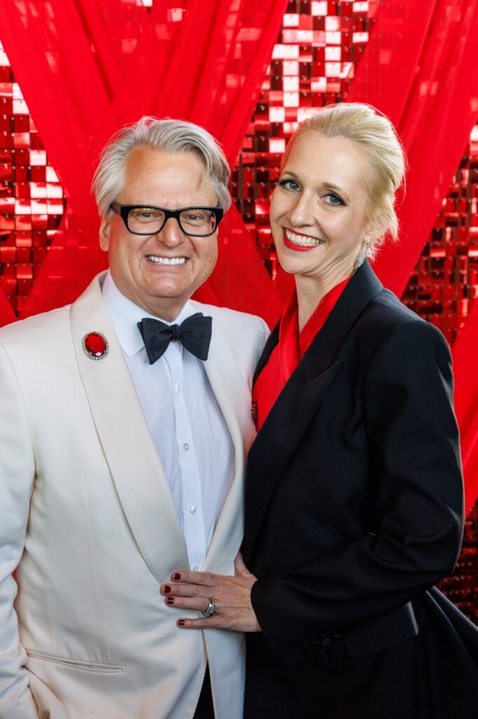 A man in a white suit jacket and black bowtie and glasses stands next to a woman in a balck suit jacket and red shirt.