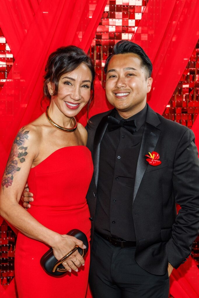 A woman in a red dress with a gold necklace stands with a man in a black suit and bowtie with a red flower in the pocket.