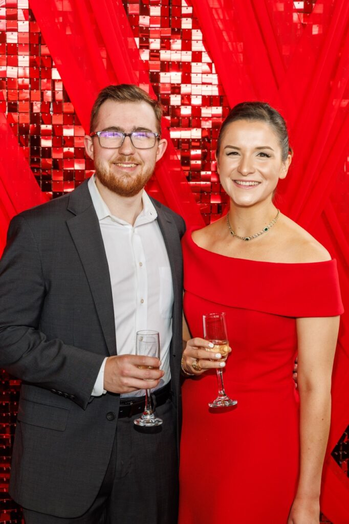 A man in a grey suit jacket and glasses holds a champagne glass with a woman in a red dress.