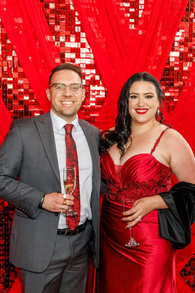 A man with a grey suit jacket and red tie stands with a woman with a red satin dress and black jacket on her arm.