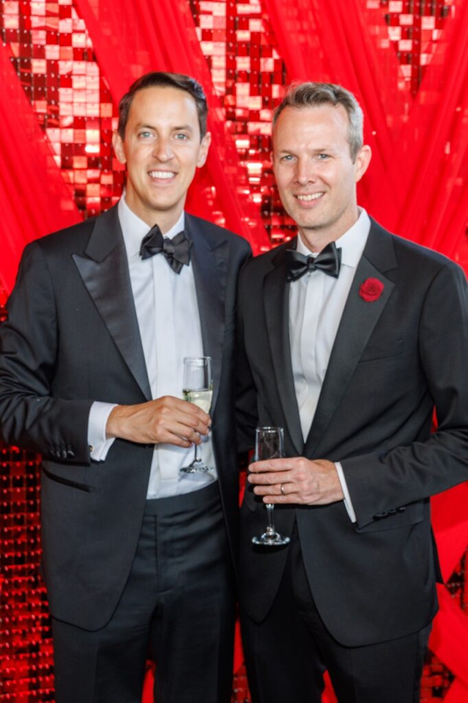 Two men in suits and bowties hold champagne glasses in front of a red background.
