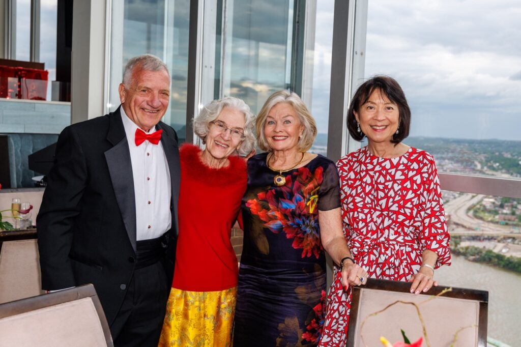 Three women and a man stand at a table in front of a window that shows the city.