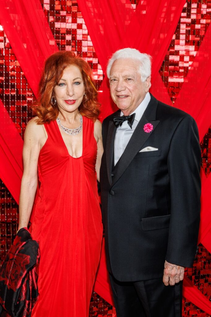 A woman in a long red dress stands with an older man in a black suit and bowtie.