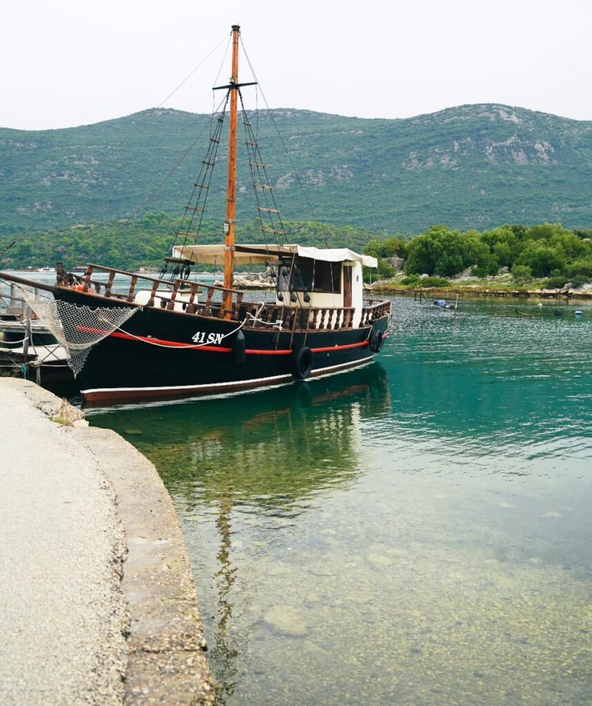 A fishing boat, black with a white cabin and a red stripe, is docked in clear blue-green water with green, tree-covered mountains in the background under a light sky.