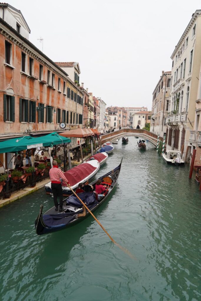 A classic Venetian gondola with a gondolier in a striped shirt and hat is seen from above, moving through a narrow canal lined with historic buildings.