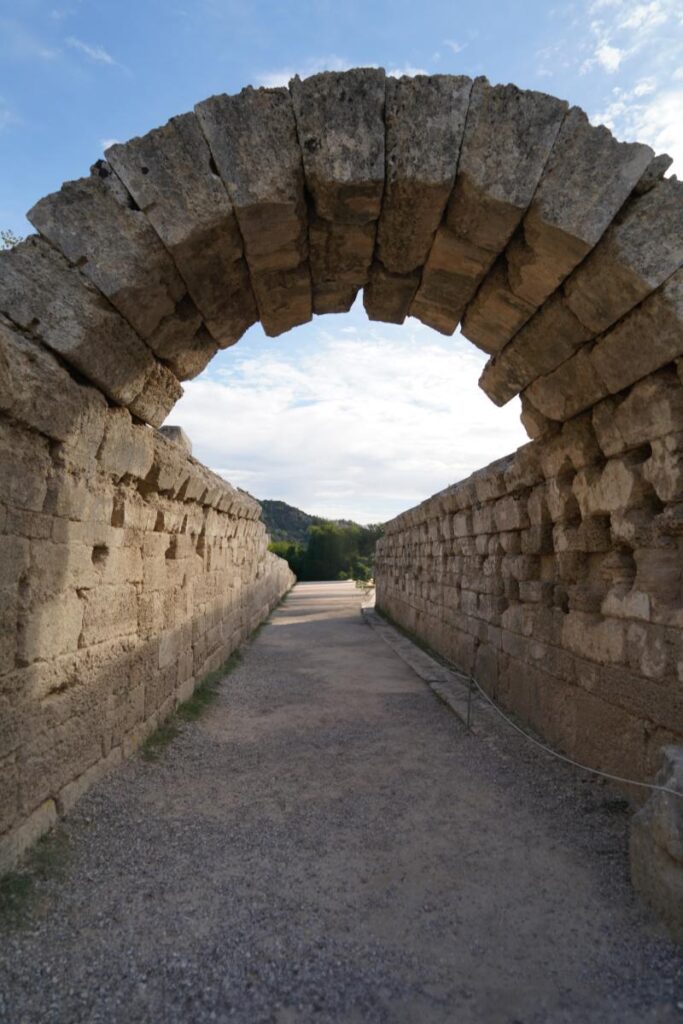 An ancient stone archway opens to a long, unpaved pathway with stone walls on either side, leading to a bright landscape under a partly cloudy sky.