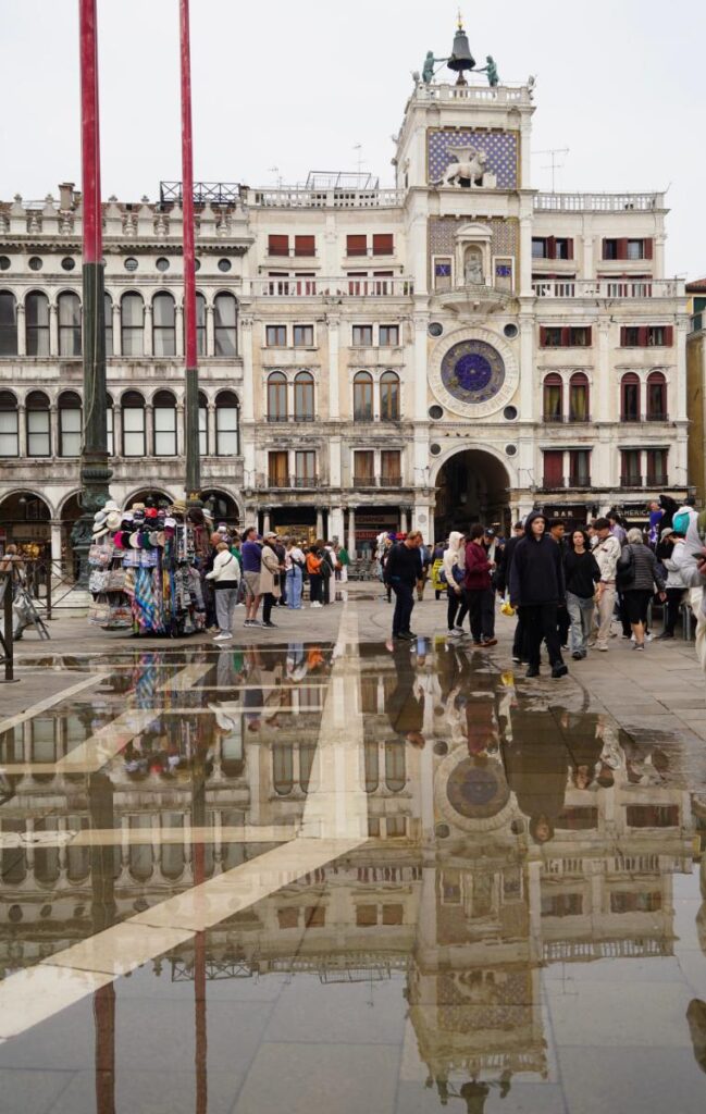 A view of St. Mark's Square in Venice, Italy, on an overcast day, showing reflections of the buildings and clock tower in puddles on the square.