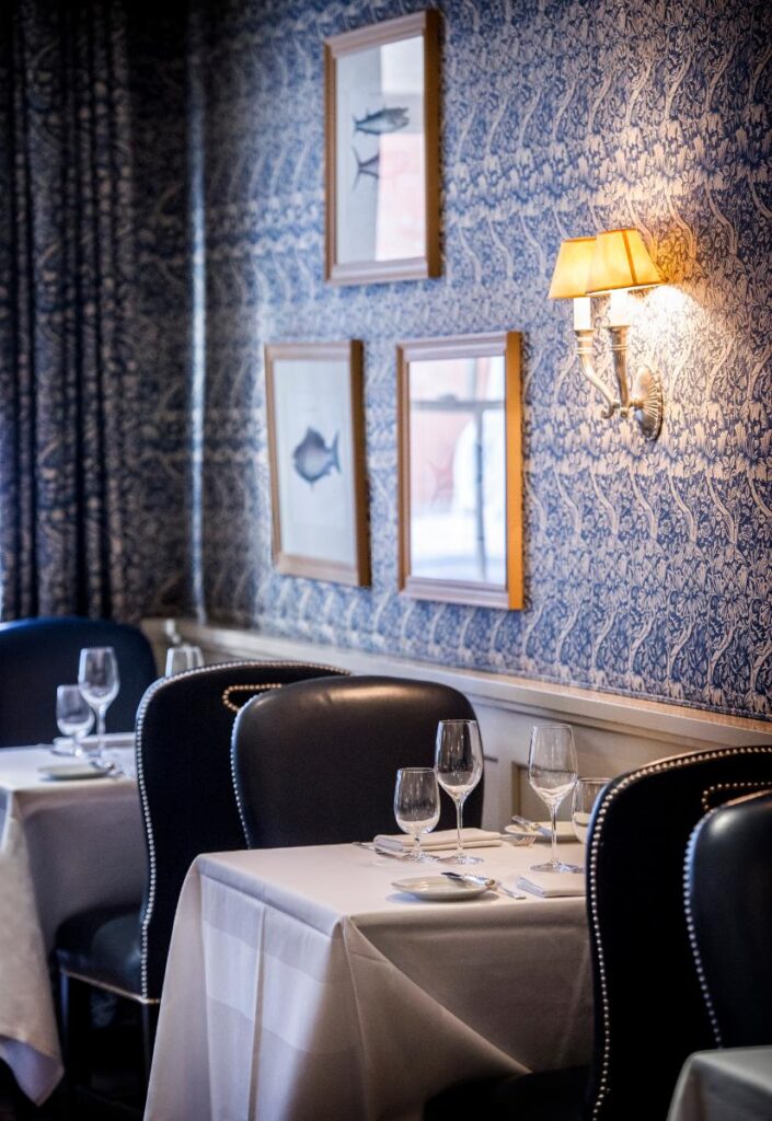 A well-lit dining room features several tables with white tablecloths and dark upholstered chairs against a wall covered in blue and white patterned wallpaper.