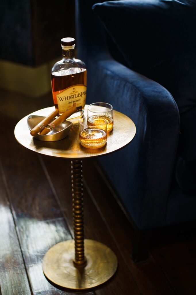 A gold little table with a bottle of whiskey and a cigar beside a blue velvet chair.