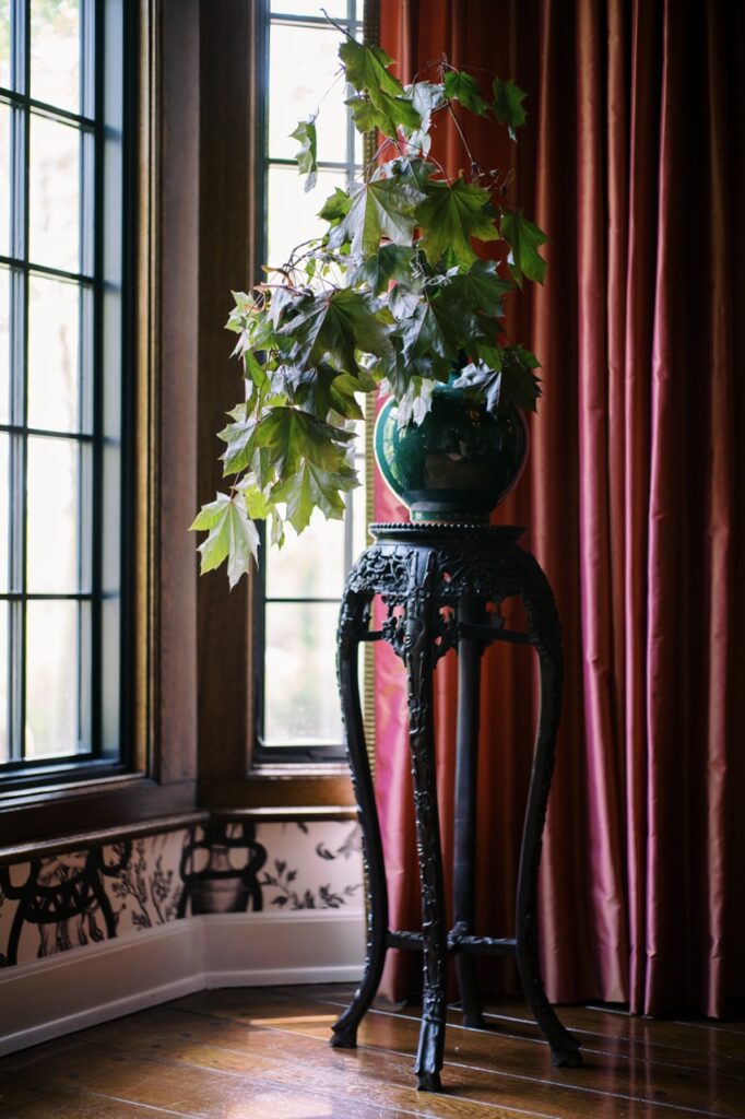 A vase stands on a tall table beside a velvet window curtain.
