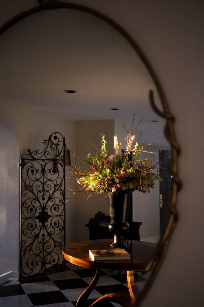A wood table with a vase and flowers reflects in an oval mirror.