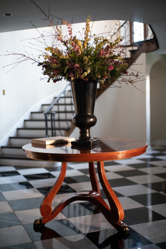 A small wooden round table on a checkered floor with a black vase and flowers on top of it.