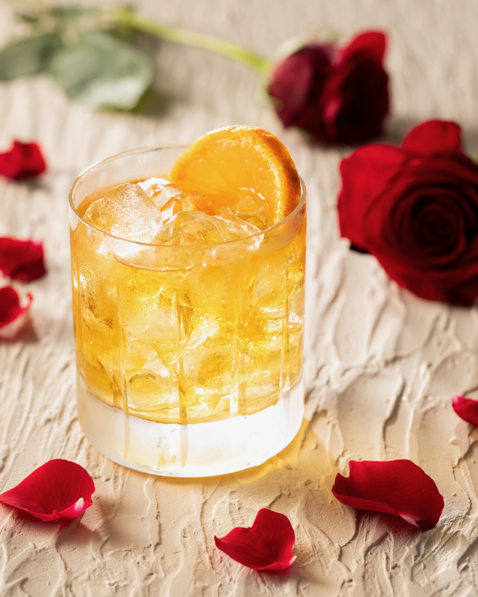 A rocks glass with ice holds an orange Moonstruck cocktail with an orange slice garnish. The glass sits amongst rose petals on a cream table.
