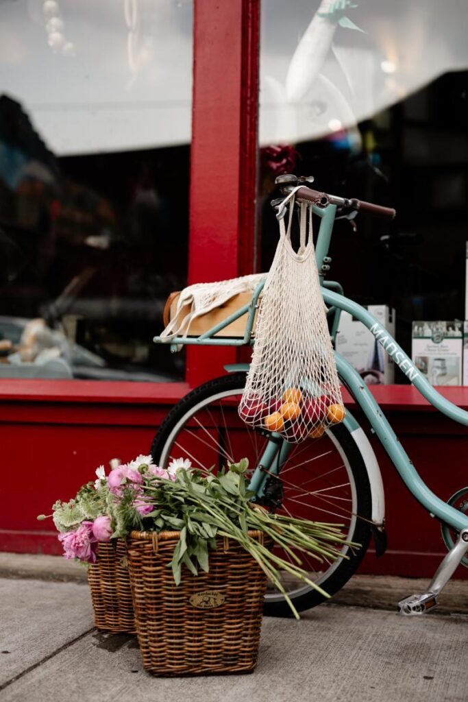 A light blue cargo bicycle is parked on a sidewalk next to a red building, with a mesh bag of oranges and apples hanging from the handlebars.