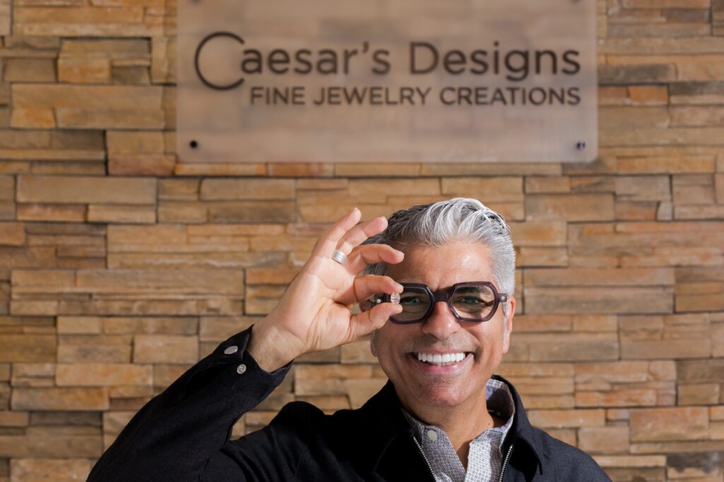 Caesar stands outside his sign holding a diamond up to his eye.