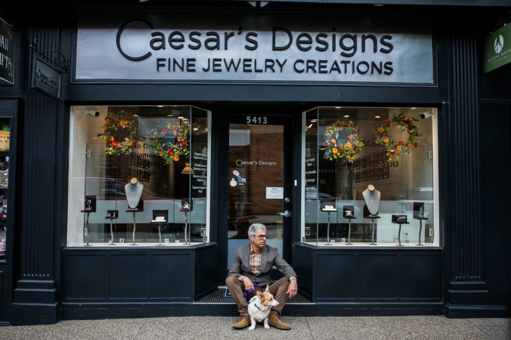 A man and his dog sit outside of Caesar's Designs building and display windows.