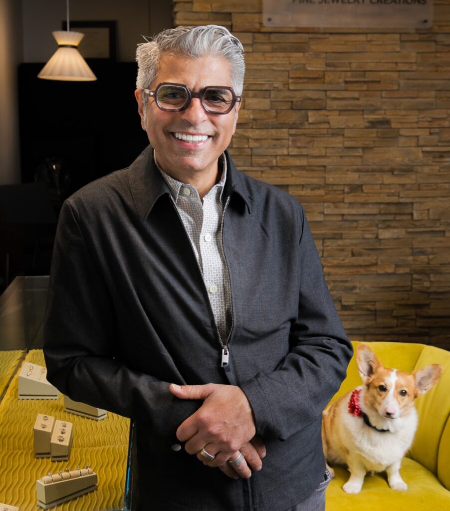 Caesar stands against a table while his dog, a corgi, sits on a yellow chair.
