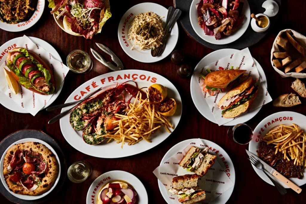 A table laden with various French dishes, including a whole lobster with fries, sandwiches, salads, and a pizza, with wine glasses and cutlery alongside.