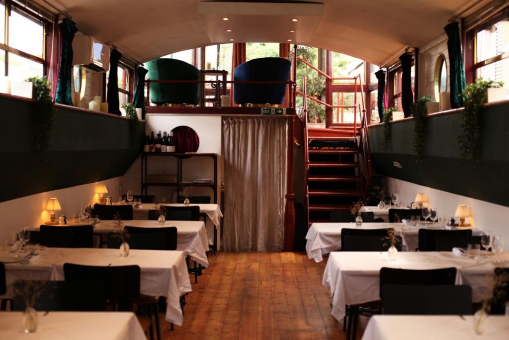 A view inside the Caravel restaurant, showing dining tables set with white tablecloths and dark chairs, leading to a staircase and an upper seating area.