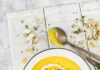 Carrot, Bell Pepper and Coconut Milk Soup An above shot of a white bowl filled with a yellow liquid, Carrot, Bell Pepper and Coconut Milk Soup, sitting on a white picnic table. A silver spoon sits to the side.