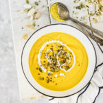 An above shot of a white bowl filled with a yellow liquid, Carrot, Bell Pepper and Coconut Milk Soup, sitting on a white picnic table. A silver spoon sits to the side.