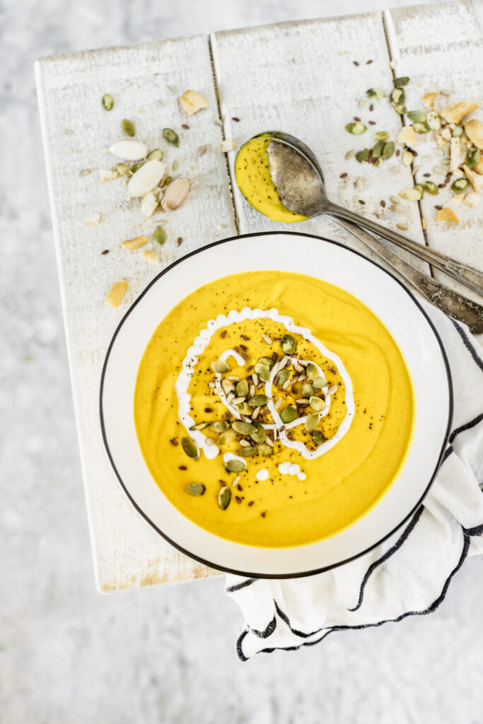 An above shot of a white bowl filled with a yellow liquid, Carrot, Bell Pepper and Coconut Milk Soup, sitting on a white picnic table. A silver spoon sits to the side.