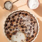 A white pie dish holds a Cherry Clafoutis with a alice taken out of it then a bowl of powder sugar and a sifter sitting above it.