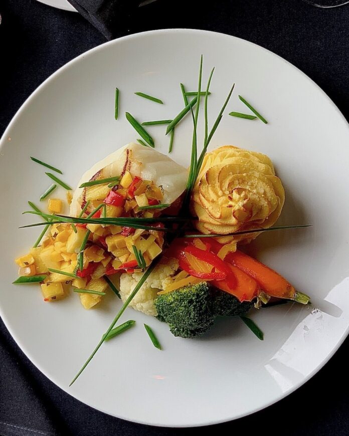 Chilean Sea Bass with a Pineapple Mango Chutney sits on a white plate with two sprigs of herbs on top of it.