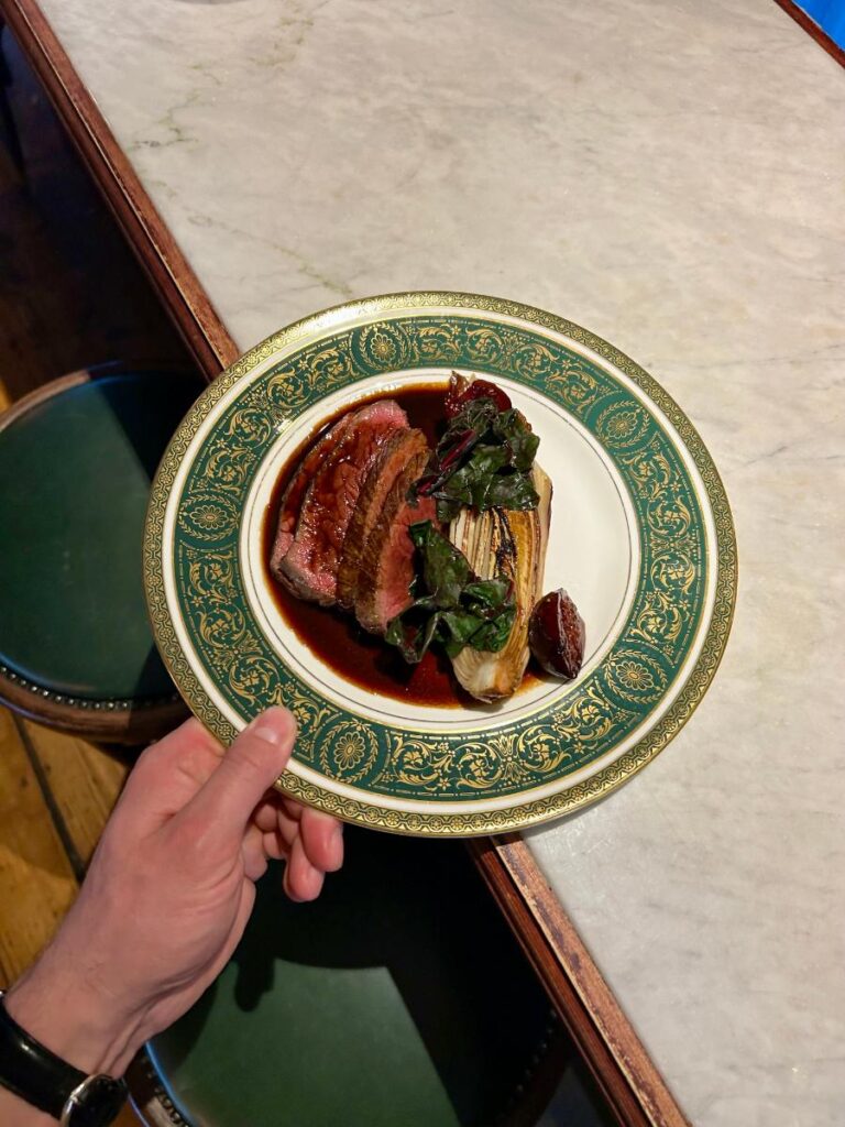 A person's hand holds a plate with sliced meat and vegetables, with a green and gold patterned rim, on a marble tabletop.
