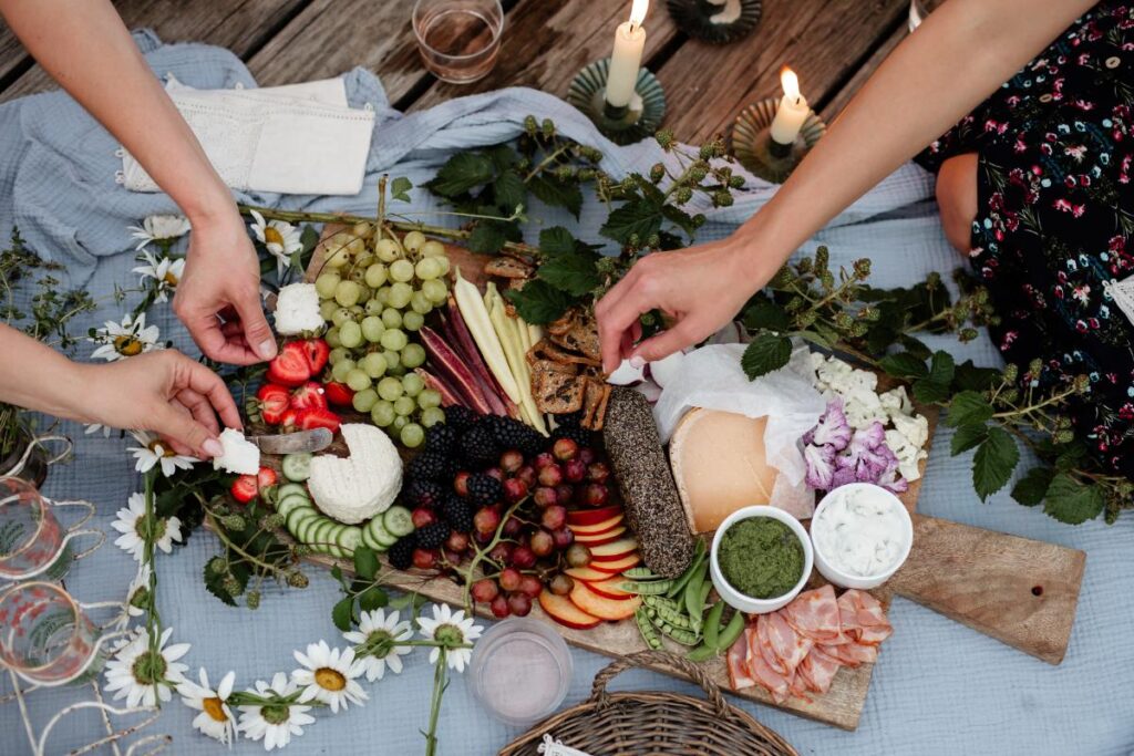 An overhead view shows two sets of hands arranging a colorful charcuterie board with grapes, berries, cheese, crackers, and cured meats on a light blue blanket.