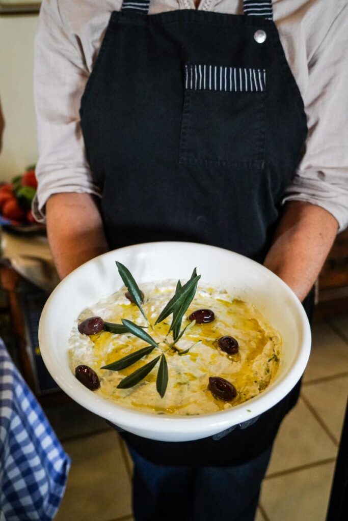 A person holds a white bowl filled with a creamy dish, possibly a dip or soup, garnished with dark olives and green leaves, with a drizzle of olive oil on top.