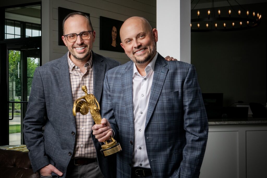 Two brothers in suits jackets stand side by side in Cranberry Dental Studio holding an award in the one on the right's hand.
