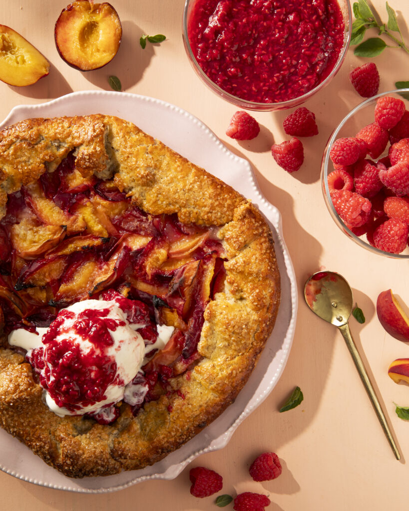 To the left side of the photo sits an Stone Fruit Galette, we see it from above, and it holds a scoop of vanilla ice cream on top.