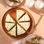 An above shot of a Zucchini and Carrot Olive Oil Cake. The cake is covered in white icing and cut into eight pieces. One slice is removed and sitting on a plate off to the top left.