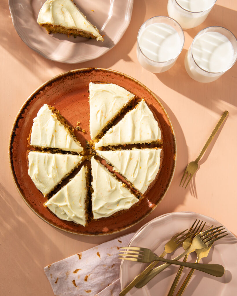 An above shot of a Zucchini and Carrot Olive Oil Cake. The cake is covered in white icing and cut into eight pieces. One slice is removed and sitting on a plate off to the top left.