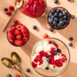 An aerial shot of Raspberry Compote with Homemade Vanilla Ice Cream in a glass bowl. Three bowls of berries sit nearby.