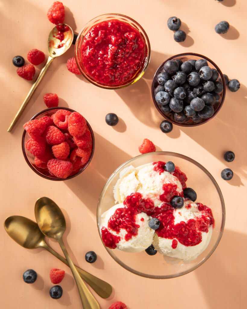 An aerial shot of Raspberry Compote with Homemade Vanilla Ice Cream in a glass bowl. Three bowls of berries sit nearby.