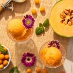 An aerial view of Husk Cherry and Melon Sorbet, an orange ice cream, scooped into two glass containers. Half a melon sits off to the side.
