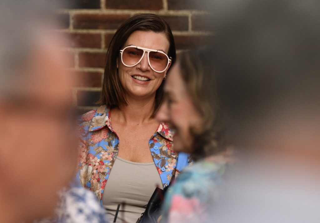A woman stands against a brick wall with large, white frame sunglasses on.
