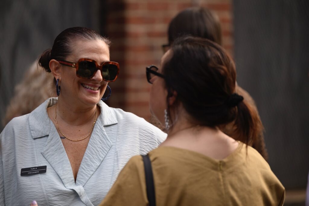 A woman talks to another woman, both have their hair tied back.