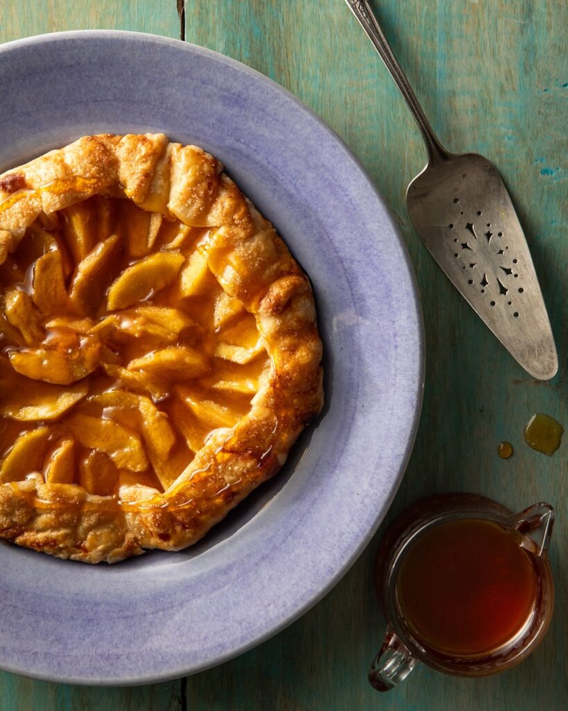 A round Easy Hot Honey Peach Galette sits on a blue plate with a pie spatula sitting beside it.