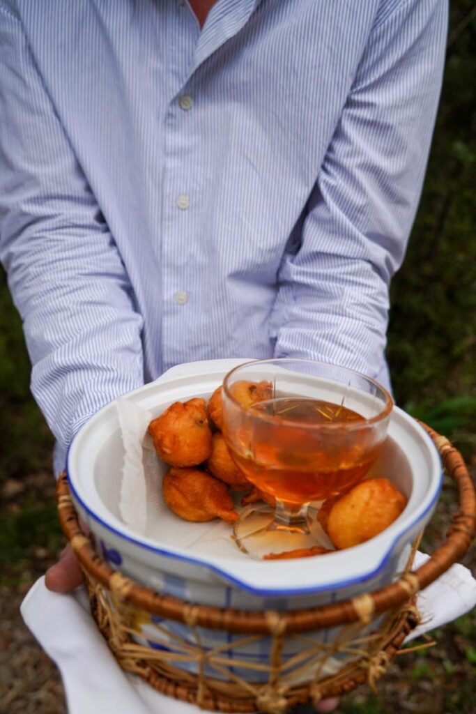 A person holds a plate of small, fried, golden-brown spheres with a glass of amber liquid in the center.